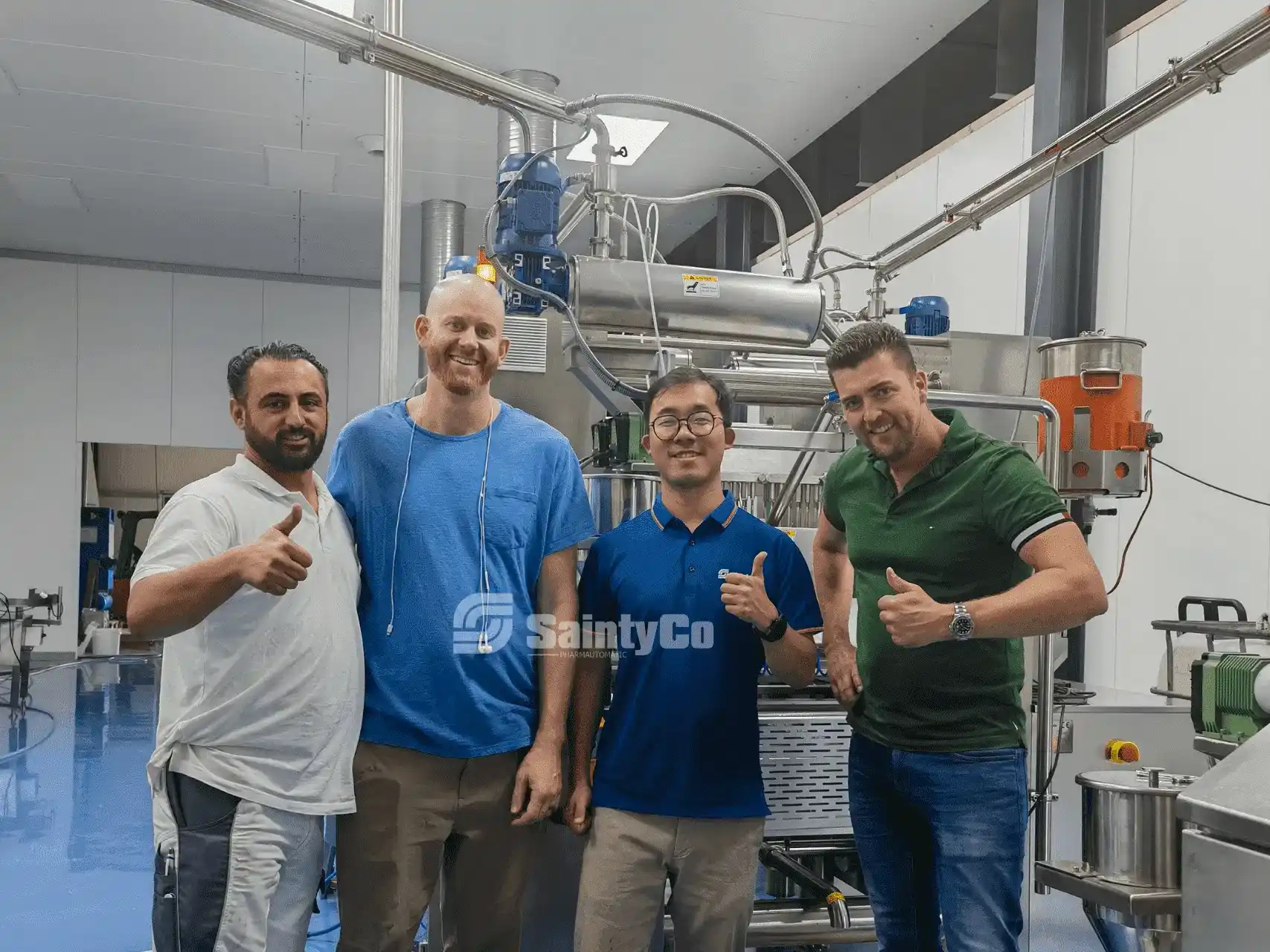 Four men stand together in a factory setting, all giving thumbs-up gestures and smiling. Positioned in front of industrial equipment, two men are wearing casual t-shirts, while the other two are dressed in polo shirts. The floor is blue, surrounded by gummy making machines from Gummygenix.