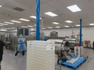 A worker in protective gear stands in a clean, industrial facility. The worker is holding an item and is near large, stainless steel machinery. Stacks of white trays are piled nearby. Blue pillars, fluorescent lights, and various equipment are visible in the background.