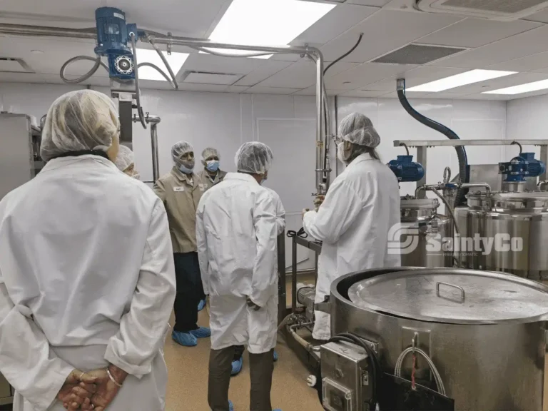 A group of people wearing lab coats, hairnets, and masks gather in a pharmaceutical manufacturing facility. They are surrounded by various stainless steel equipment and machinery, including a gummy making machine. The logo "SaintyCo" is visible on one of the machines.