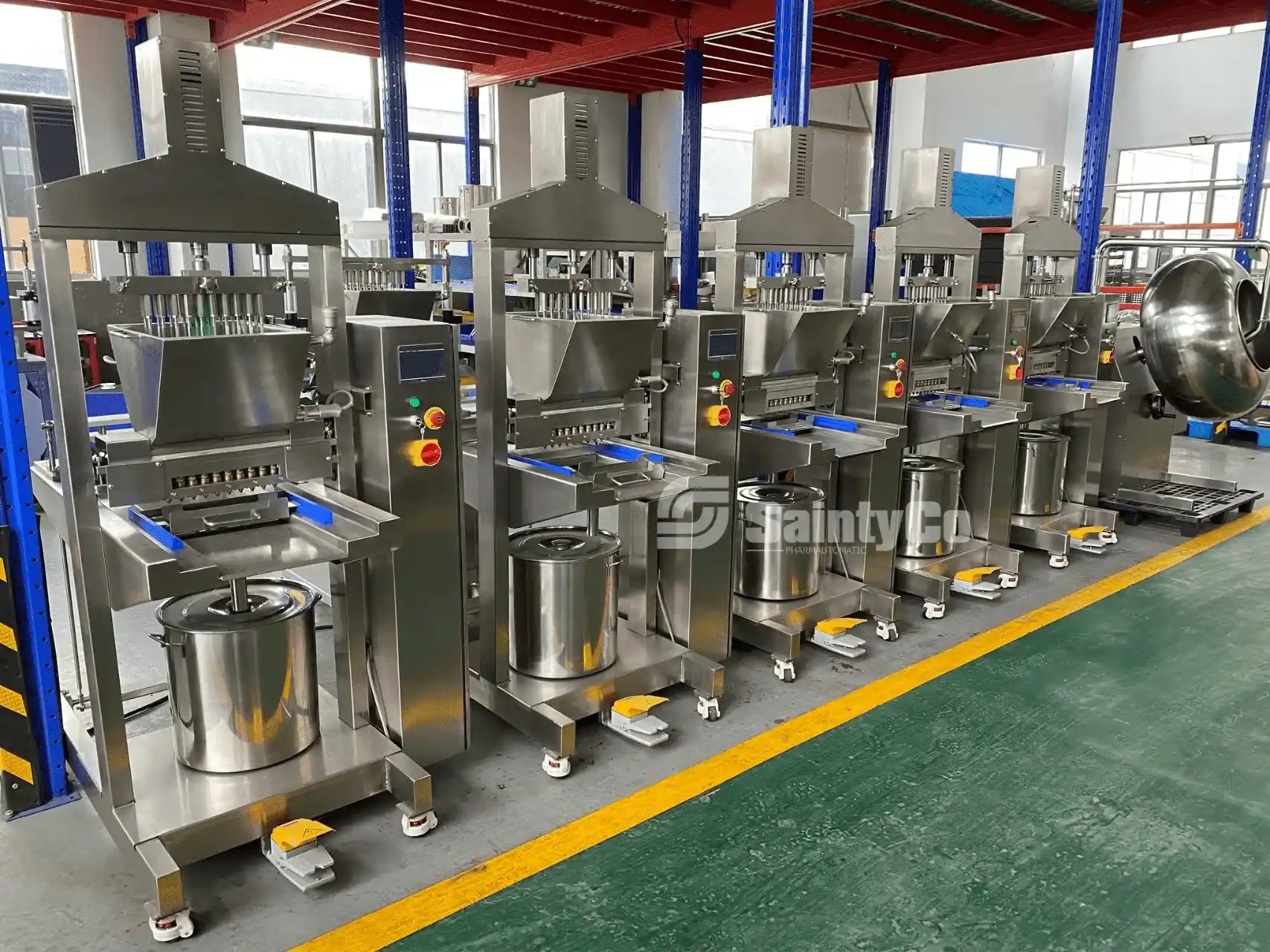 A row of industrial-grade stainless steel food processing machines lined up in a factory setting. Each machine has control panels with buttons, various compartments, and containers for processing materials. The floor is green with yellow safety lines.