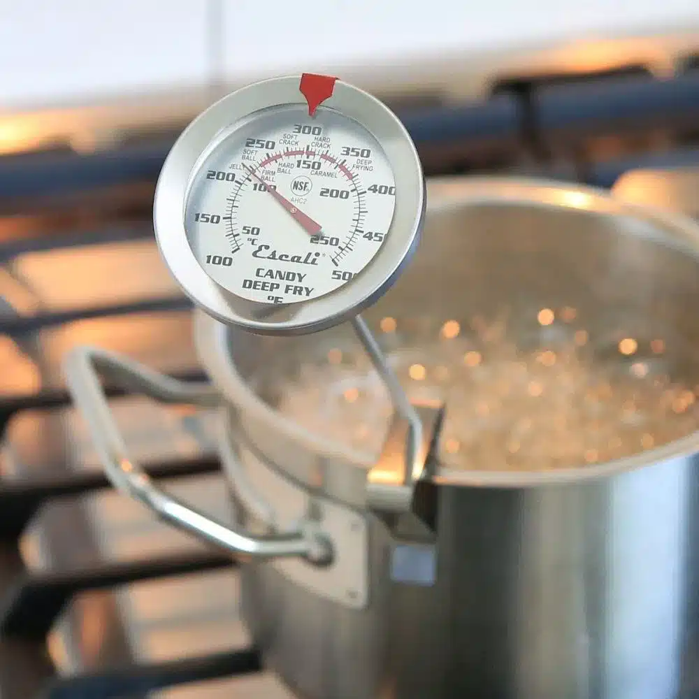 A metal pot on the stovetop contains bubbling liquid, essential in the gummy manufacturing process. A candy and deep fry thermometer is clipped to the pot, displaying a temperature range from 100 to 400 degrees Fahrenheit. The stovetop burners are visible in the background.