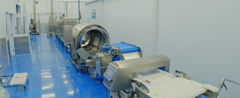 A modern, clean industrial facility featuring a row of stainless steel food processing machines on a glossy blue floor. The setup includes automated conveyors and inspection systems beneath bright overhead lighting, alongside the advanced SaintyCo GummyGenix Sugar Coating Drum for gummies production.