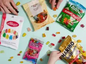 A variety of gummy candies are displayed on a light blue surface. Five packages, showcasing different gummy bear brands and flavors, are opened, including Albanese, Trader Joe's, Black Forest, Project 7, and Haribo. Hands eagerly reach for the assorted sweets scattered around.