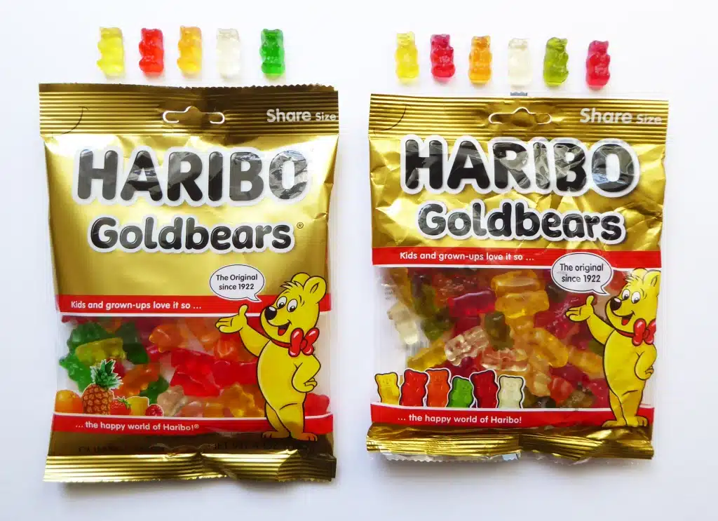 Two packets of Haribo Goldbears gummy candy are displayed. One is unopened, showcasing its vibrant packaging with colorful gummy bears and the iconic Haribo logo, while the other is open with candies arranged in a single line above, highlighting the variety of flavors offered by this beloved gummy bear brand.