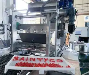 A tablet press machine operates in a manufacturing facility. Red tablets are visible on a tray labeled "SAINTYCO" in the foreground. The machinery is metallic with various components and controls.