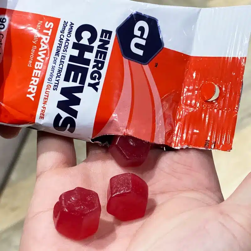 A hand holding three red hexagonal energy chews next to an open GU Energy Chews packet labeled "Strawberry.