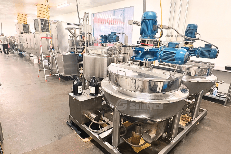 Industrial kitchen with stainless steel gummy making machinery from GummyGenix by SaintyCo, including large mixing bowls and motors, lined up in a clean, spacious room. Ingredients and equipment are organized along the counter. People work in the background.