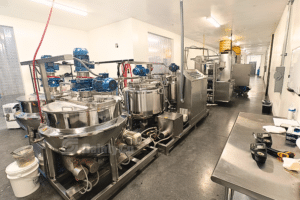 A commercial gummy production kitchen with large stainless steel vats and mixing equipment lining the walls, supplied by SaintyCo. The room is well-lit with fluorescent lights, and various controls and tools are visible on the tables. The floor is dark and polished.