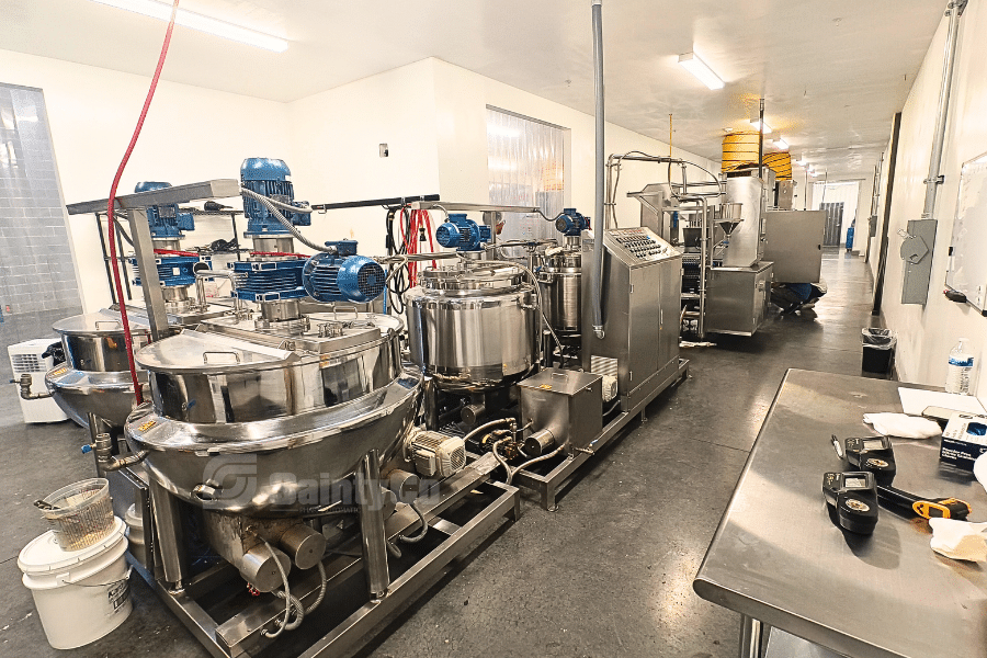 A commercial gummy production kitchen with large stainless steel vats and mixing equipment lining the walls, supplied by SaintyCo. The room is well-lit with fluorescent lights, and various controls and tools are visible on the tables. The floor is dark and polished.