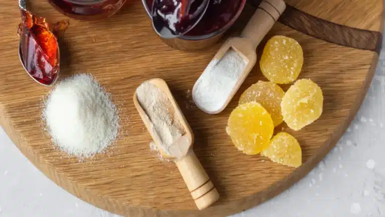 A wooden board displays ingredients including a mound of white powder, a scoop of beige powder, a scoop of white powder, several round yellow sugar-coated candies, and jars of red jam. A spoon with orange jam rests nearby.