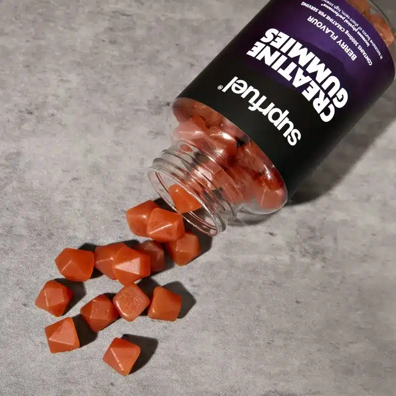 A tipped-over bottle of berry-flavored creatine gummies spills out onto a gray surface. The red, pyramid-shaped creatine gummies scatter around the open bottle, showcasing the precision of production machinery’s handiwork.