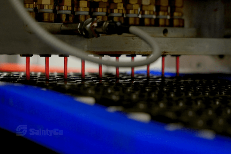 Close-up of a gummy mold filling machine in operation. Red nozzles are dispensing material into gummy molds on a moving conveyor. The background includes metal components and a blue surface.