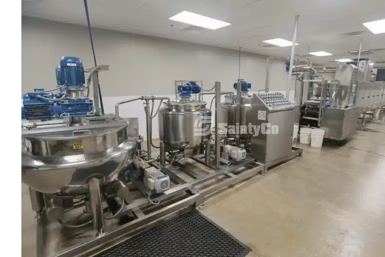 Industrial gummy processing equipment with stainless steel vats and control panels in a clean facility. The machinery includes blue motorized components and interconnected pipes, designed for manufacturing or chemical processing. The floor is polished concrete.