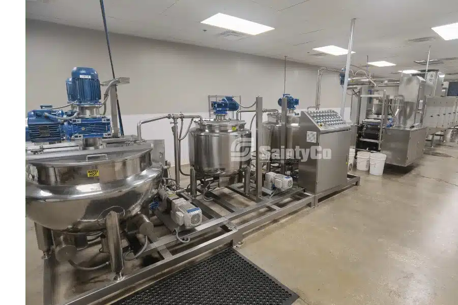 Industrial gummy processing equipment with stainless steel vats and control panels in a clean facility. The machinery includes blue motorized components and interconnected pipes, designed for manufacturing or chemical processing. The floor is polished concrete.