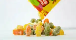 A colorful pile of gummy bears is being poured out from a partially visible yellow candy bag. The bears are in various colors, including orange, yellow, green, and red, against a plain white background.