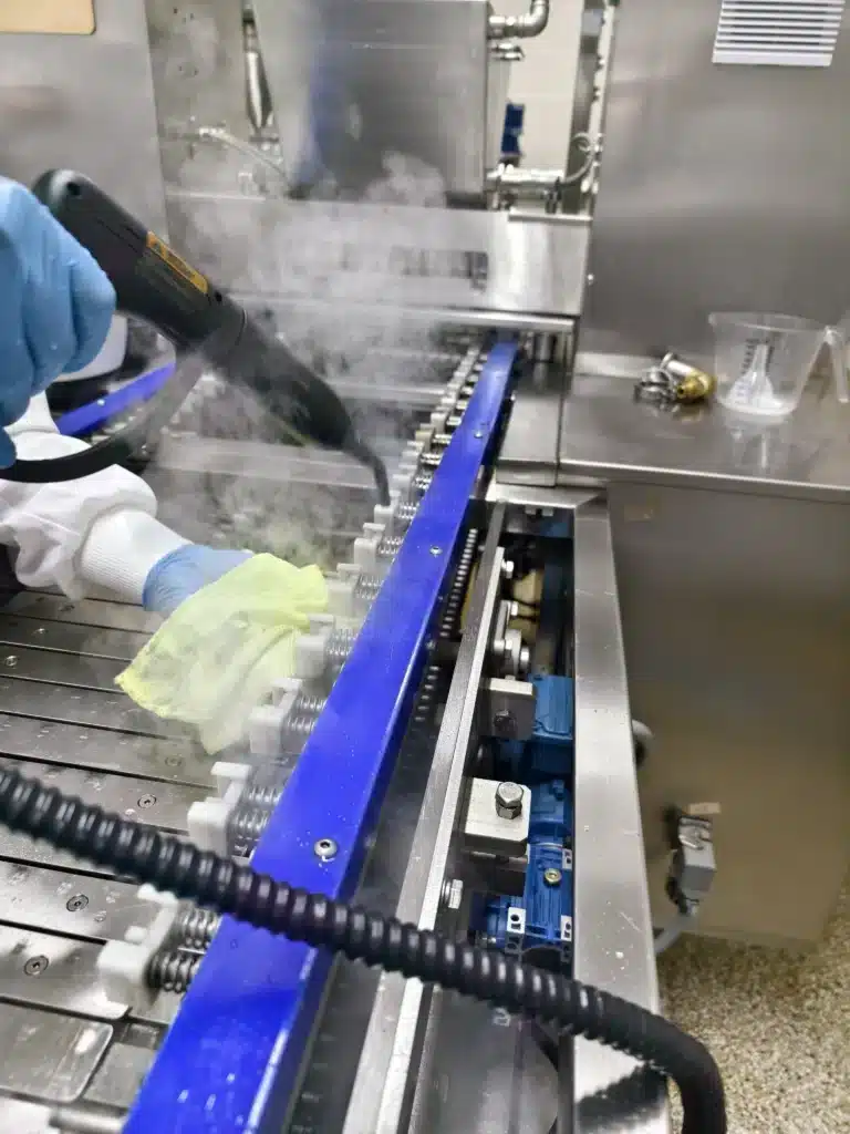 A person in protective clothing and gloves uses a handheld steam cleaner on an industrial conveyor belt. Steam is visible, and there are metal and plastic components around the machine, with a measuring cup nearby.