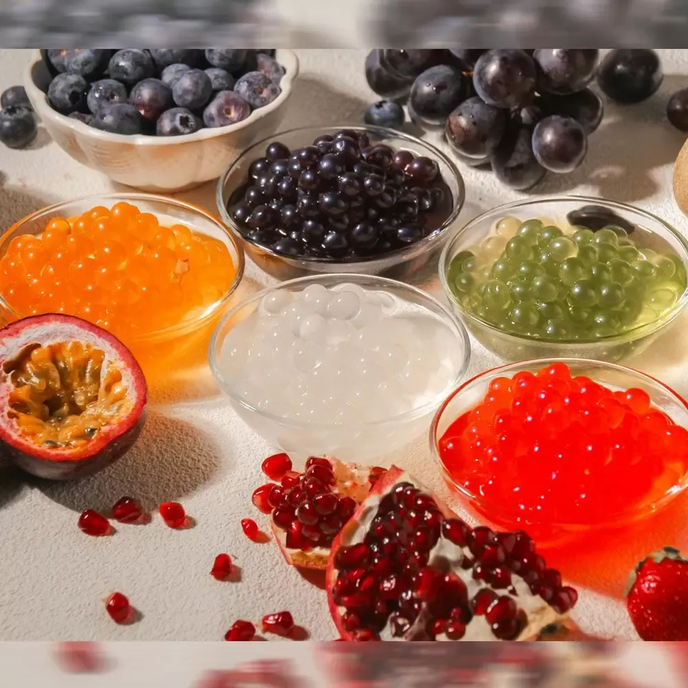 A variety of colorful popping boba balls in small bowls are arranged with fresh fruits like blueberries, purple grapes, passion fruit, pomegranate, and strawberries on a white surface. The boba colors include orange, black, green, clear, and red.