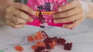 Hands holding a pink bag of gummy candies being poured onto a marble surface. Various red and orange bear-shaped gummies are visible.