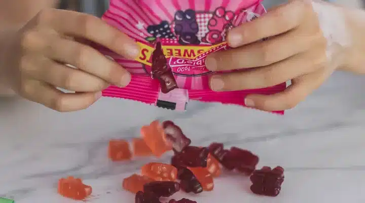 Hands holding a pink bag of gummy candies being poured onto a marble surface. Various red and orange bear-shaped gummies are visible.