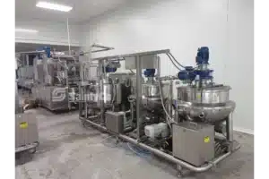 A industrial kitchen with large stainless steel machinery used for gummy processing. The SaintyCo gummy making equipment includes multiple tanks and mixers, connected with pipes and valves, set up in a clean, tiled environment.