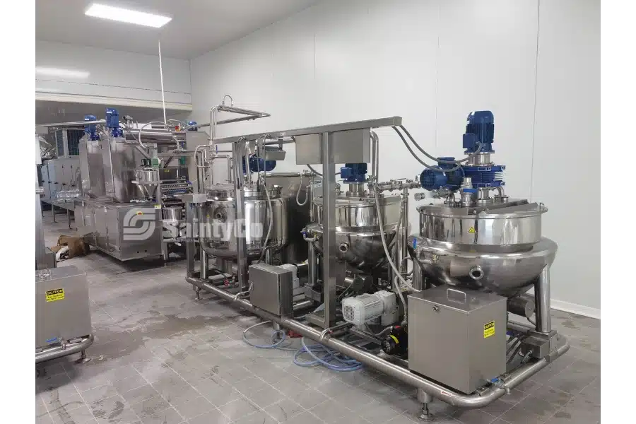 A industrial kitchen with large stainless steel machinery used for gummy processing. The SaintyCo gummy making equipment includes multiple tanks and mixers, connected with pipes and valves, set up in a clean, tiled environment.