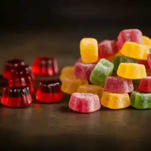 A pile of sugar-coated gummy candies in various colors, including yellow, green, pink, and orange, sits next to several red and black dome-shaped gummies on a dark surface. The background is blurred.