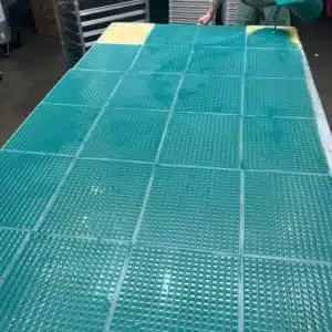 A person sanding a large work surface covered with green transparent plastic mats in a workshop setting. The person is partially visible, focusing on smoothing the edges of the mats, with additional equipment in the background.