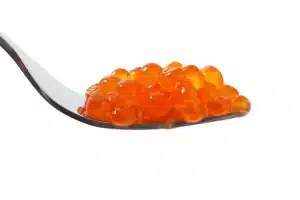 A close-up of a metal spoon holding bright orange fish roe, also known as salmon caviar, against a white background.