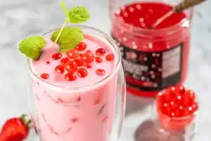 A glass of pink strawberry milk topped with red popping boba and a sprig of mint, with a jar of red popping boba and a strawberry in the background.