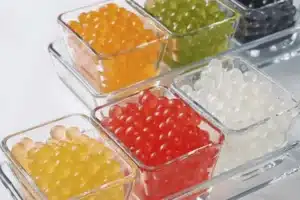 Six small glass dishes containing colorful spheres of various flavors, including yellow, orange, red, green, white, and black, arranged neatly on a clear tray against a white background.