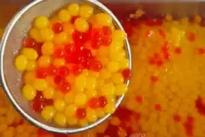 A metal strainer holding yellow and red popping boba balls over a container filled with more boba balls in liquid.