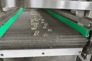 Small translucent capsules travel along a metal conveyor with green side guards, likely part of a facility using a GummyGenix popping boba maker or SaintyCo popping boba machine for efficient production.