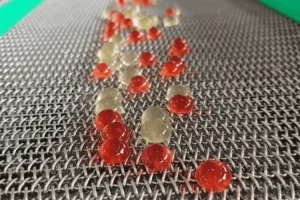 Red and clear spherical gel capsules are scattered on a metal mesh conveyor belt, likely in a facility using a GummyGenix popping boba maker or SaintyCo popping boba machine for manufacturing or processing.