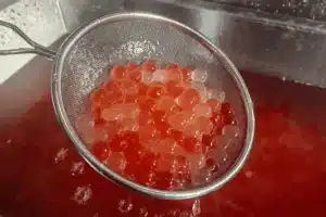 SaintyCo metal strainer holds a cluster of shiny, translucent red spheres—likely prepared using a GummyGenix popping boba maker—over a container filled with red liquid, possibly fish eggs or roe being rinsed and prepared.