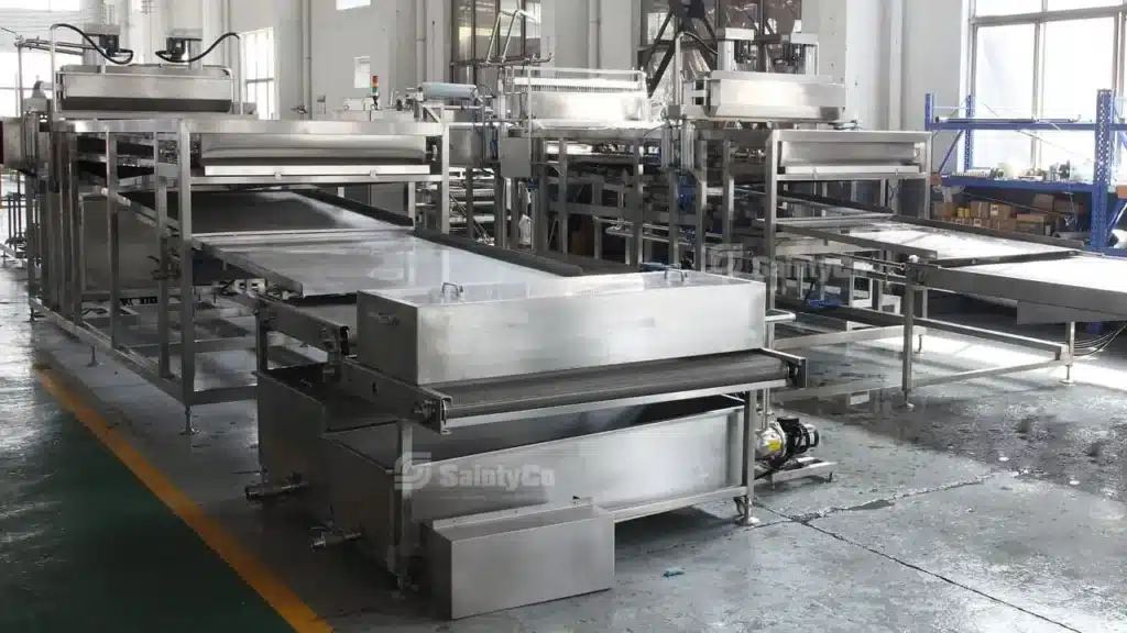 Fully automatic popping boba processing machinery and production lines of GummyGenix by saintyCo with stainless steel conveyors and components inside a factory. GummyGenix automatic popping boba machine is designed for efficient operations in food production. The factory floor is clean, with various equipment and shelves in the background.