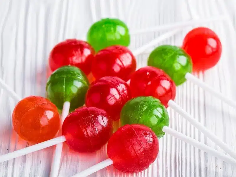 A group of red, green, and orange round lollipops with white sticks arranged on a white wooden surface.
