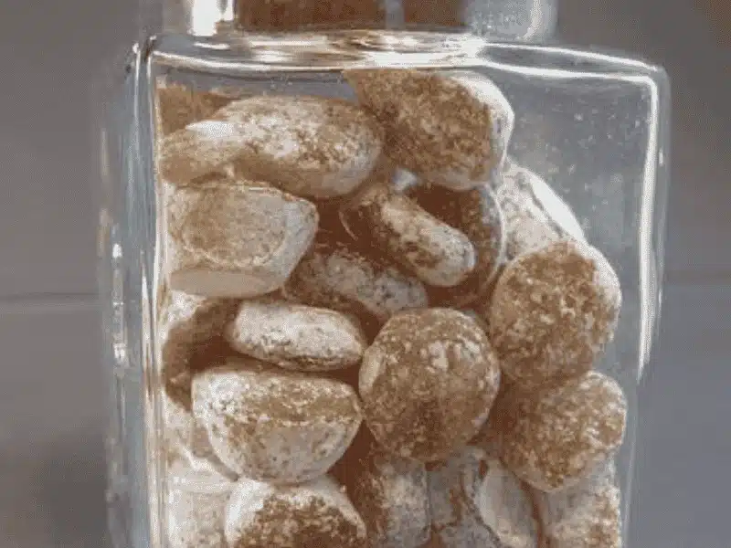 A clear glass jar filled with brown, round candies or cookies, each coated in a dusting of white powder, sits on a gray surface.