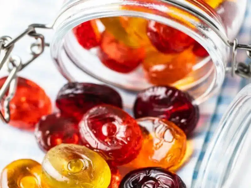 A glass jar tipped over, spilling colorful hard candies in red, orange, and yellow onto a white and blue checkered surface.