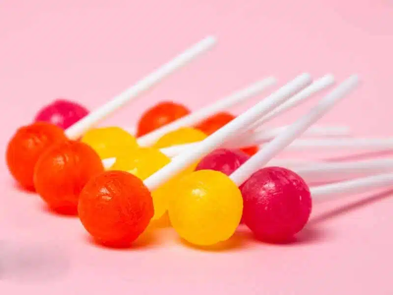 Colorful lollipops with white sticks, including orange, yellow, and pink candies, are arranged in two rows on a light pink background.