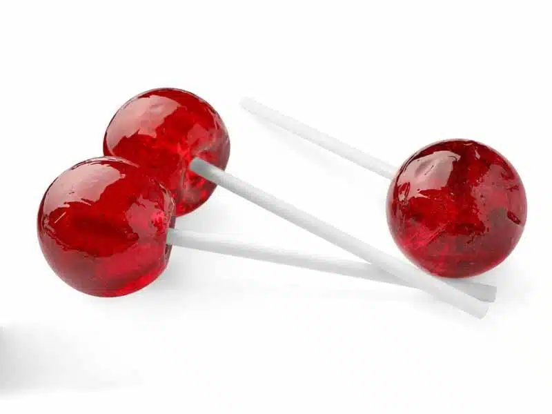 Three red lollipops with white sticks are lying on a white surface. The candies are shiny and round, reflecting light.