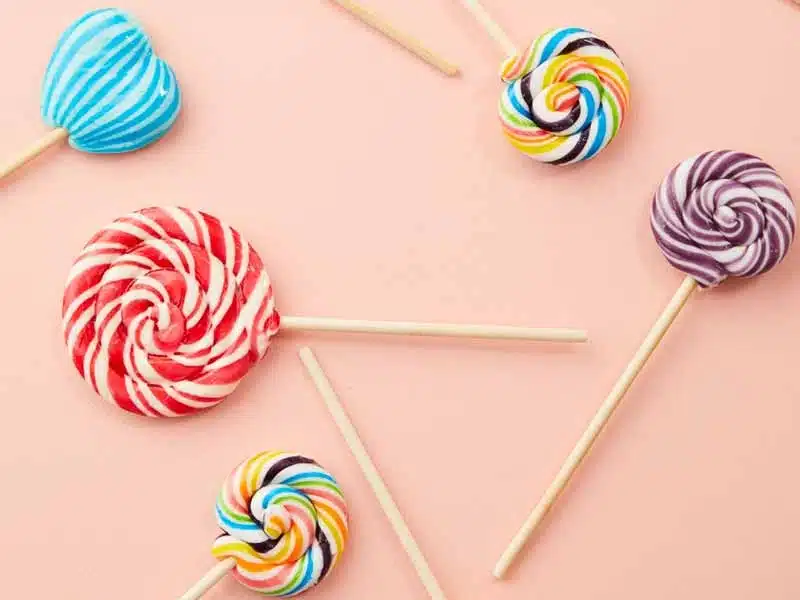 Colorful lollipops in various shapes and swirled patterns, including heart and round shapes, arranged on a light pink background. Some lollipops are rainbow-colored, while others are red, purple, or blue.